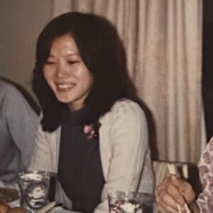 Linda sitting at a dinner table, smiling towards a glass with Santa Claus etched on it. Her shoulder-length black hair wisps over a pink flower brooch. A black blouse and white sweater fashionably juxtapose.