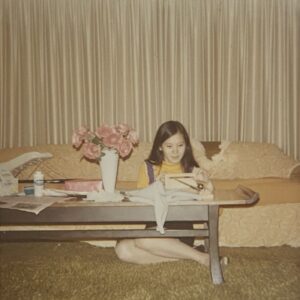 Linda shines in a mustard yellow shirt and blue tank dress while sitting at a coffee table in front of a pale yellow couch. In the center of the cluttered tabletop is a vase of pink roses. The beige drapes and brown carpet fill the rest of the frame.